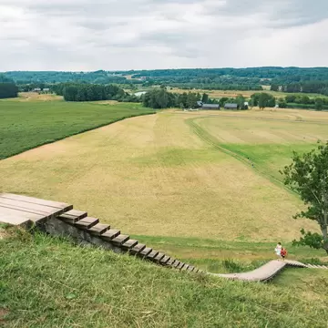 Wooden steps descend a grassy hill fort at Kernavė toward wide green fields and forest in the Neris valley.