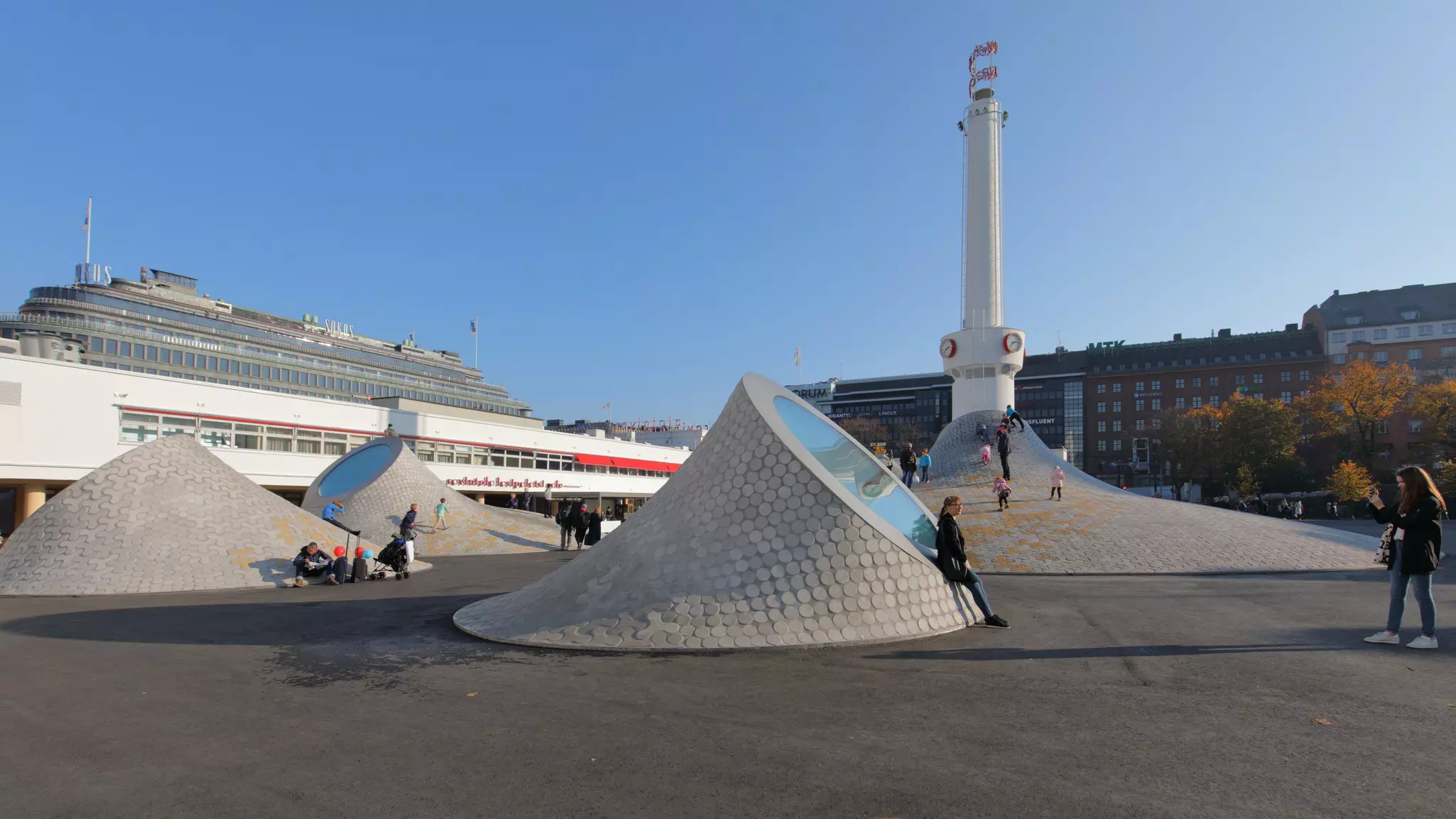 Tiled, interestingly shaped buildings each have a circle of reflective glass; people walk on them and lean against them.