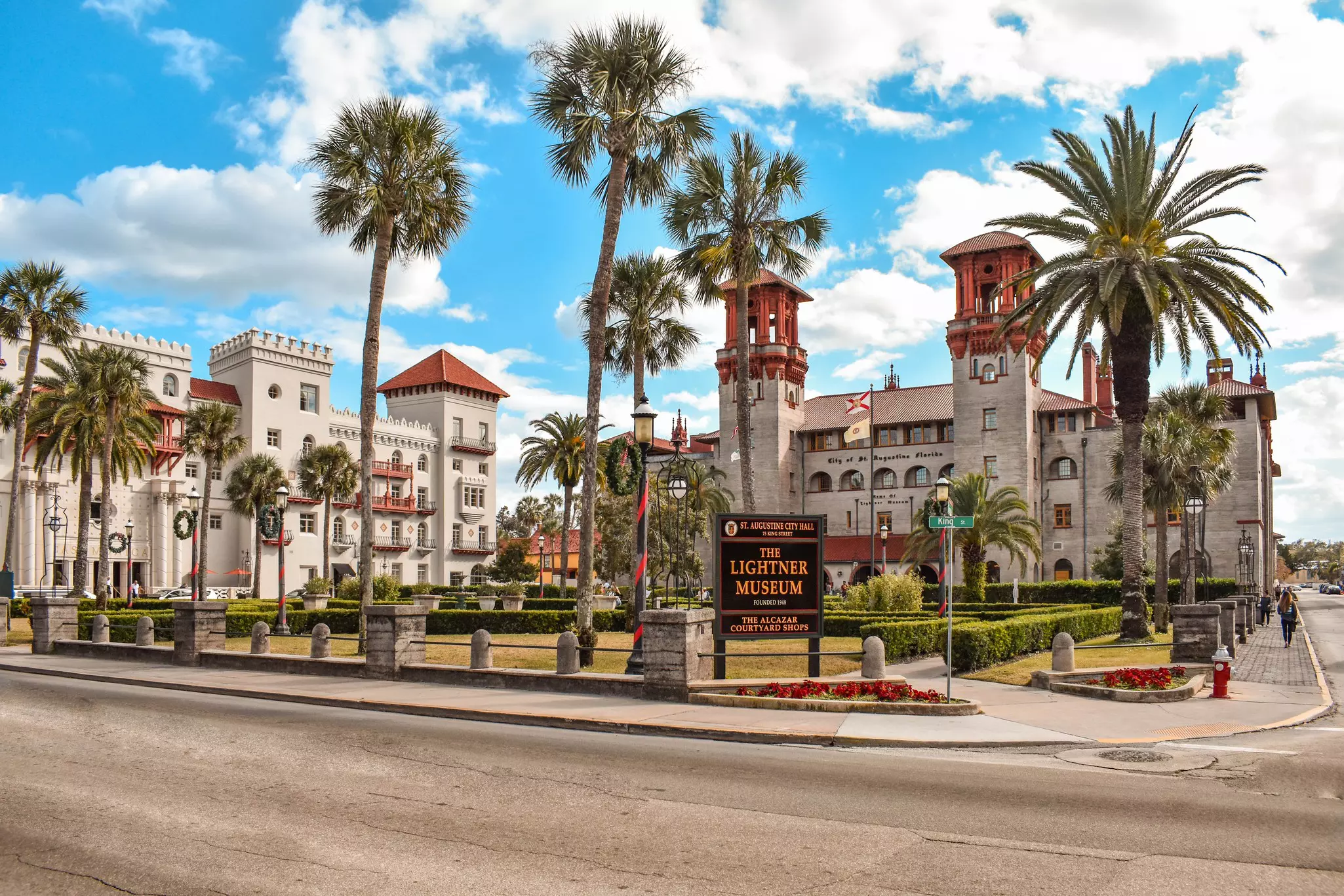 A large building with two turrets set in gardens dotted with palm trees. A sign outside indicates it is the Lightner Museum.