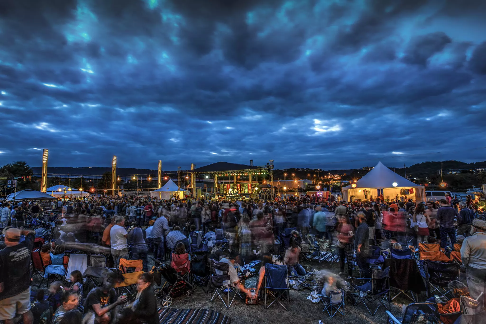Concert crowd sat on the ground at an outdoor music festival with large dark clouds in the sky above the stage