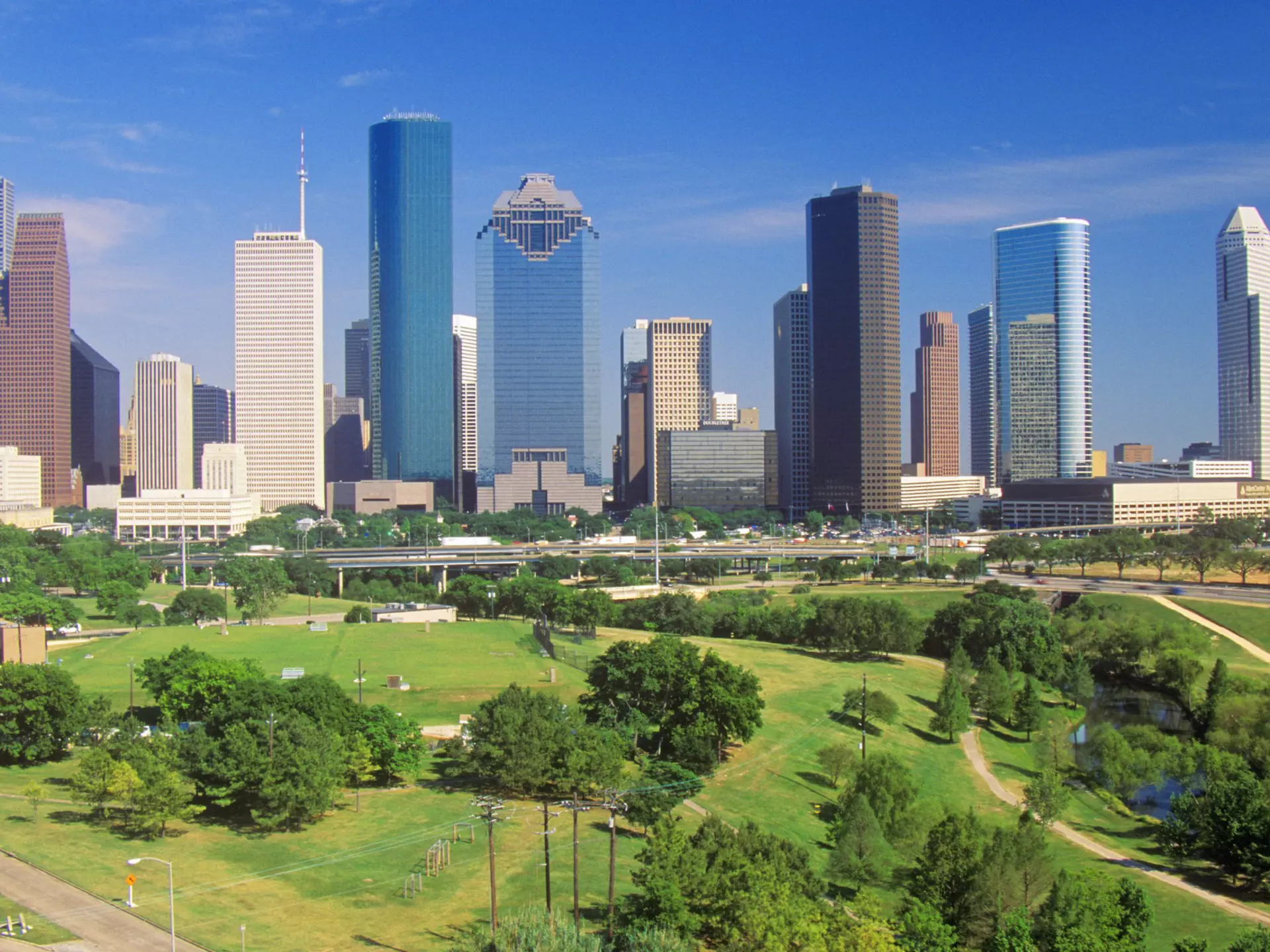 A distant view of Houston's skyline in the afternoon with Memorial Park in the foreground. The park is an expansive carpet of green, dotted with trees.