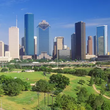 A distant view of Houston's skyline in the afternoon with Memorial Park in the foreground. The park is an expansive carpet of green, dotted with trees.