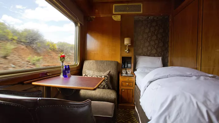 A single bed in a wood-panelled room with a seat and table next to a large window.