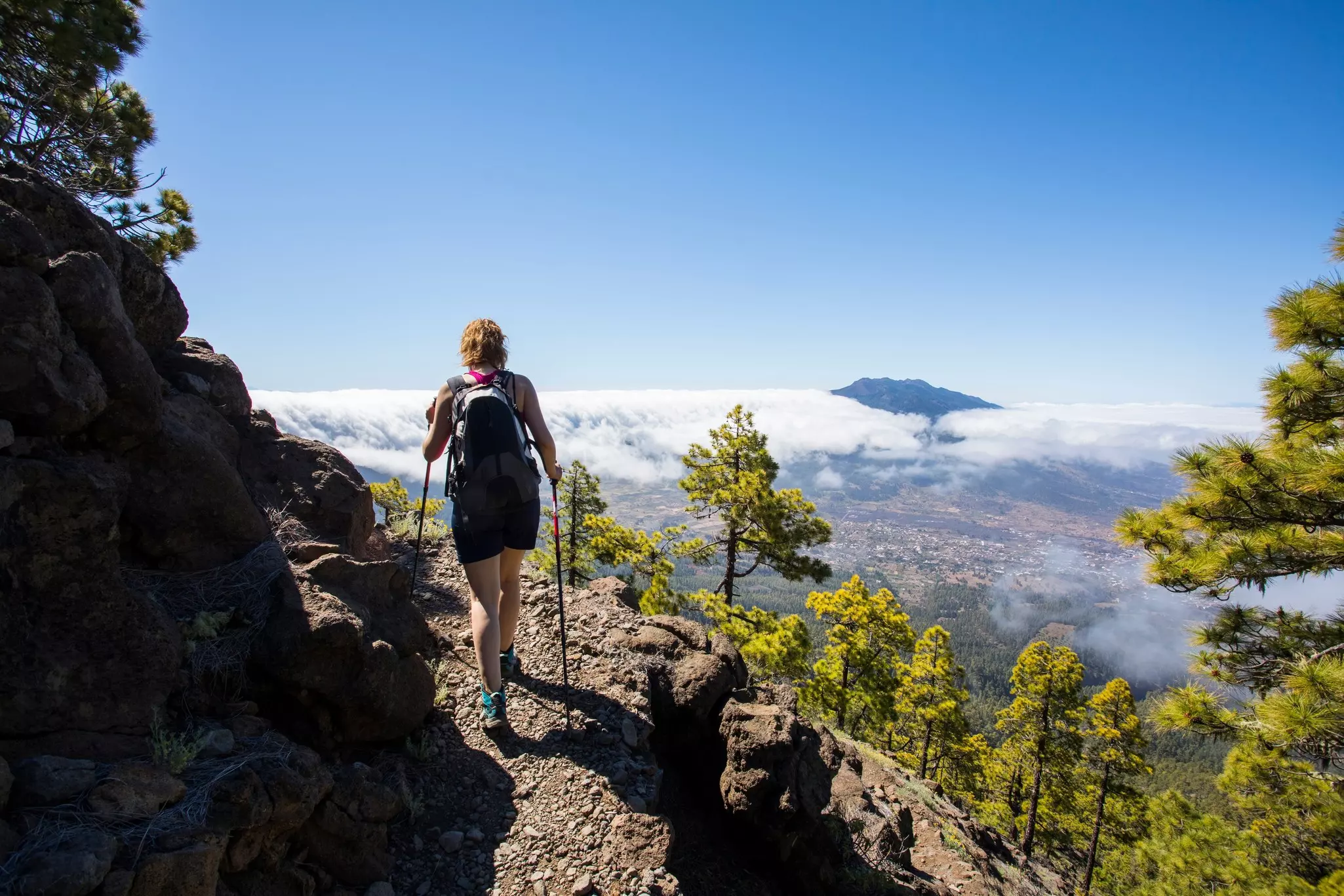 A female hiker with walking poles hikes on a narrow path on the side of a mountain with a view of trees and clouds in the distance