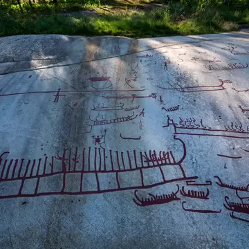Smooth pale rock slab covered in red-painted outlines of long boats, people and animals, forest at the top.