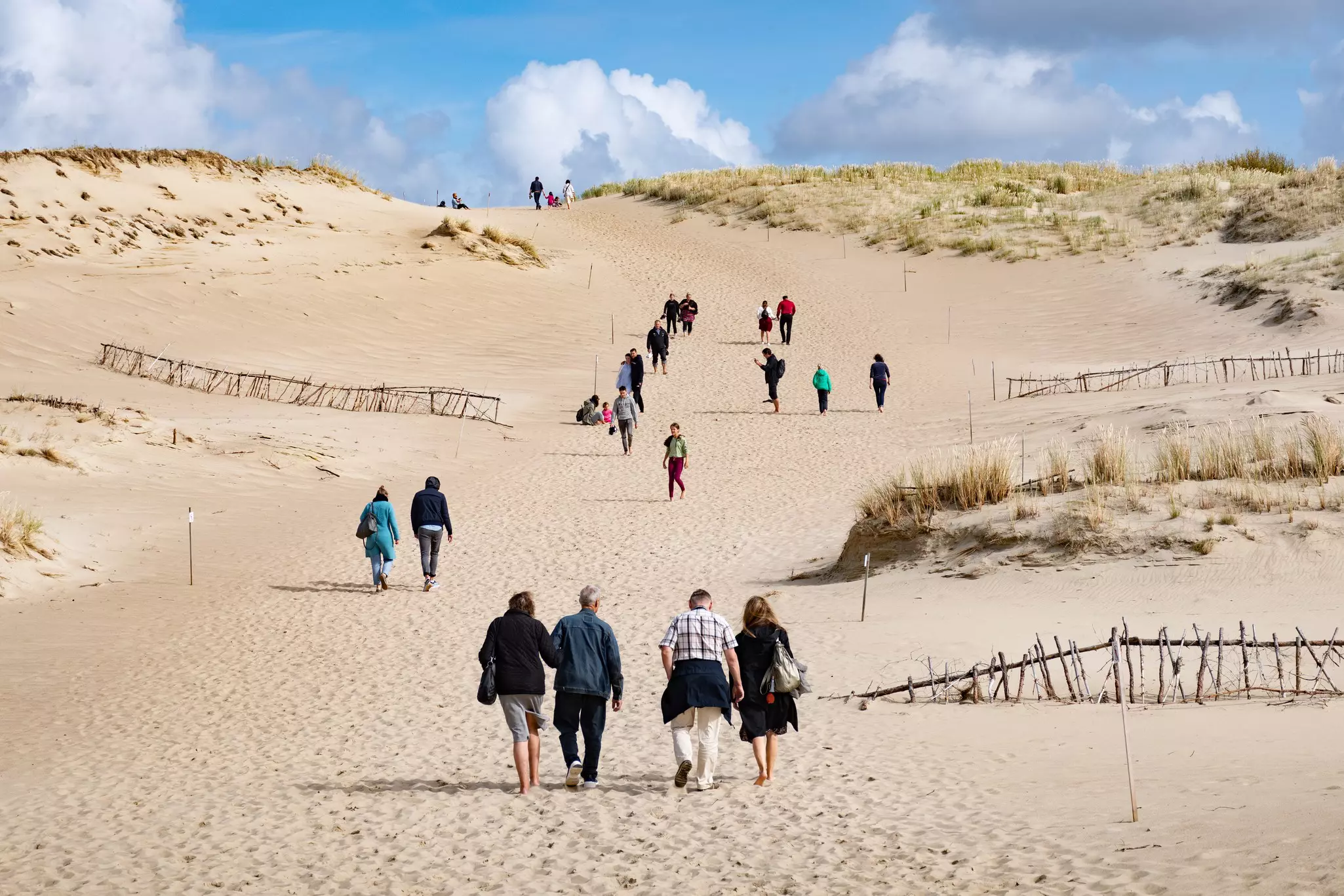 A wide shot of people walking on dunes. Partially submerged fences emerge from the sand