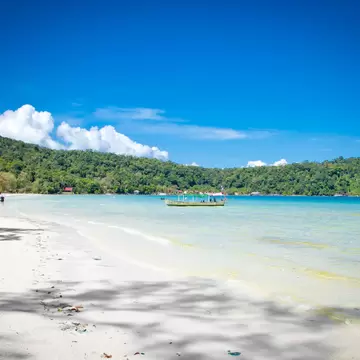 Long white-sand beach with a few people walking, clear turquoise shallows, a moored boat and green forested hills.