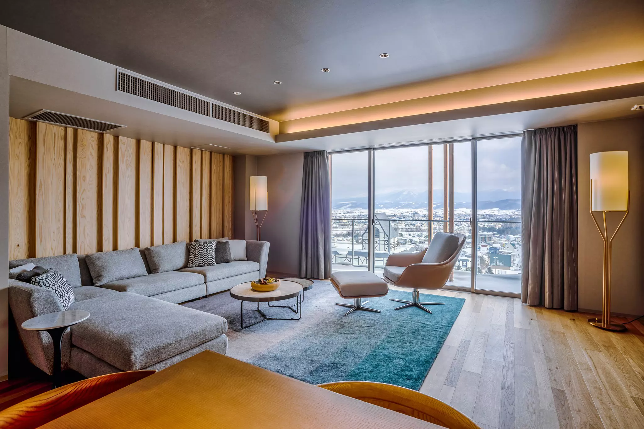 A large hotel room with a corner sofa in a lounge area facing a floor-to-ceiling window with city and mountain views.