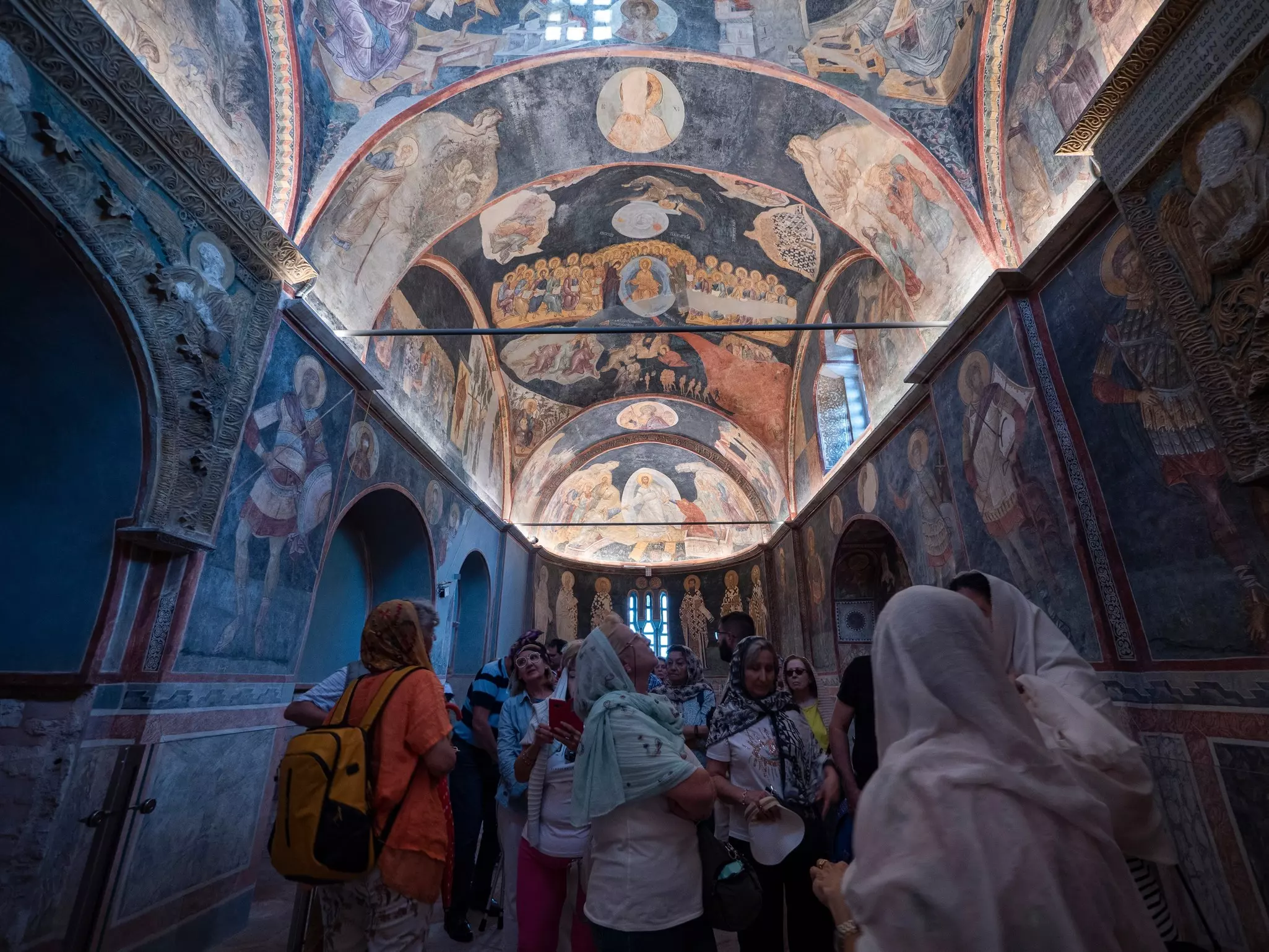 People dressed modestly with their heads covered gaze at mosaics and frescoes on the walls and ceiling of a room with a curved ceiling