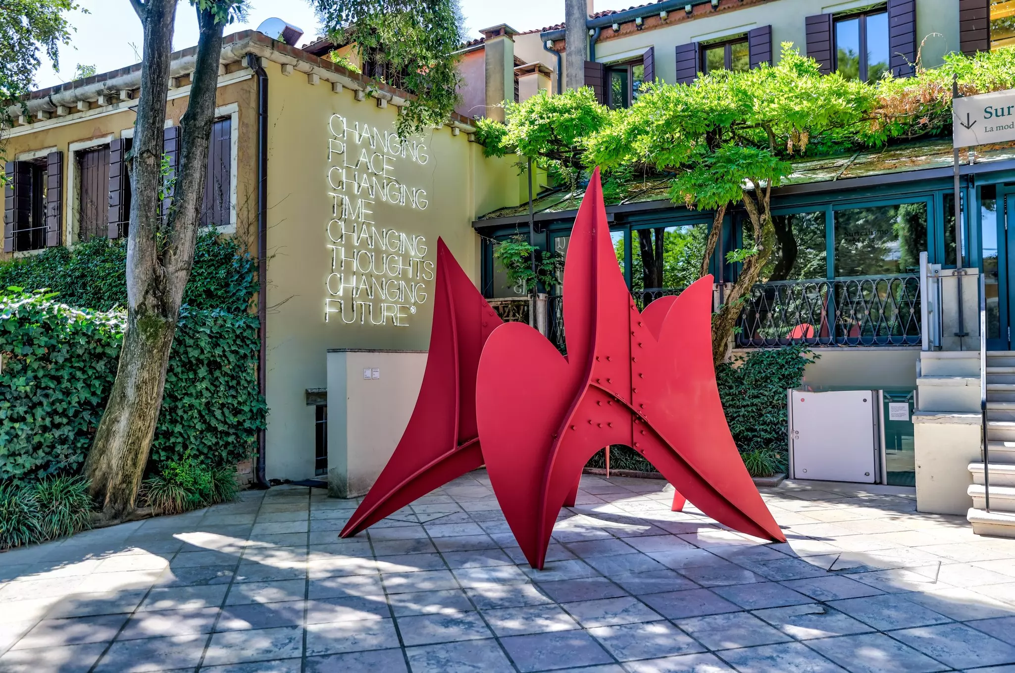 A red metal sculpture is on view in a courtyard with trees and ivy. Another work of written text is displayed on the wall of the courtyard.