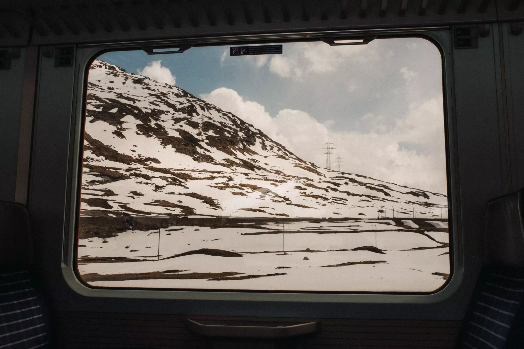 A view of a snowy peak through a train car window with clouds and blue sky.