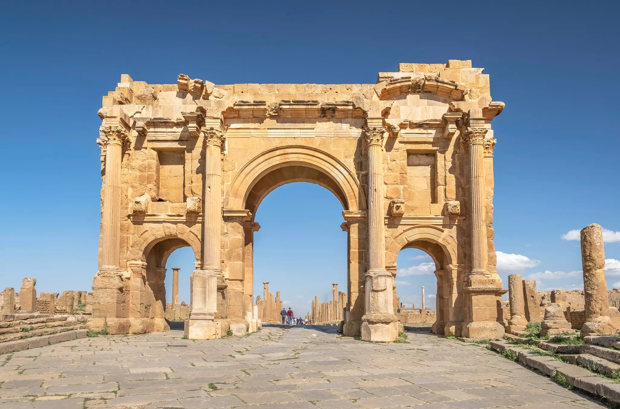An intact Roman arch of golden stone with three openings against blue sky; visible in the distance through the arch, visitors walk on a path through columns.