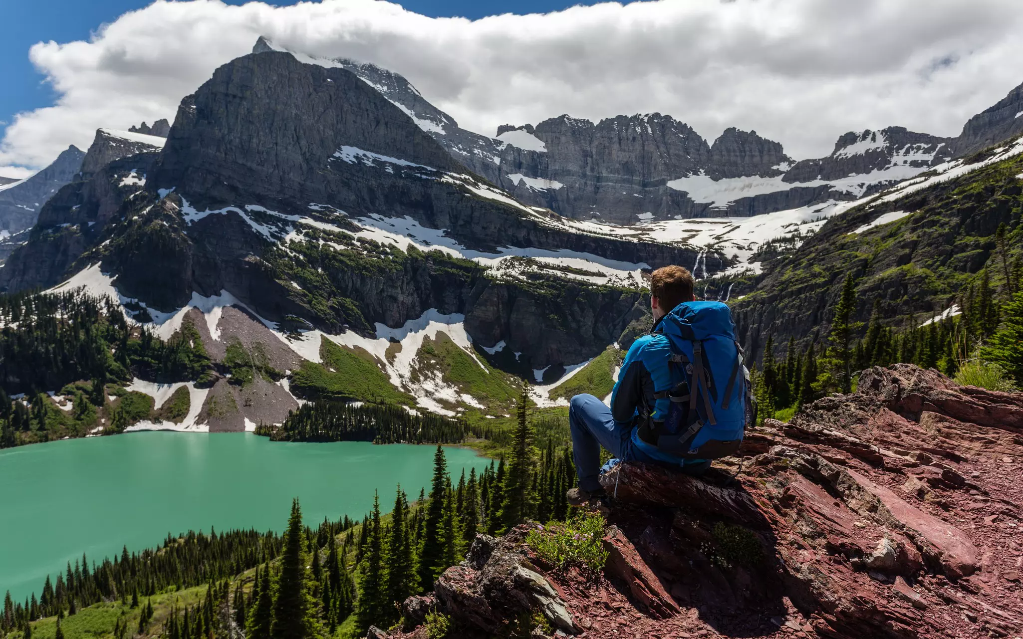 A hiker sitting on reddish rocks looking at a calm green lake and snowy mountain.