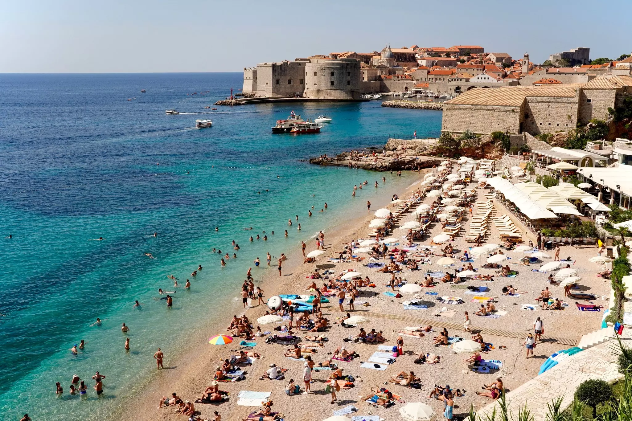A crowded sandy beach by turquoise water; a fortress at the edge of Dubrovnik is in the background.