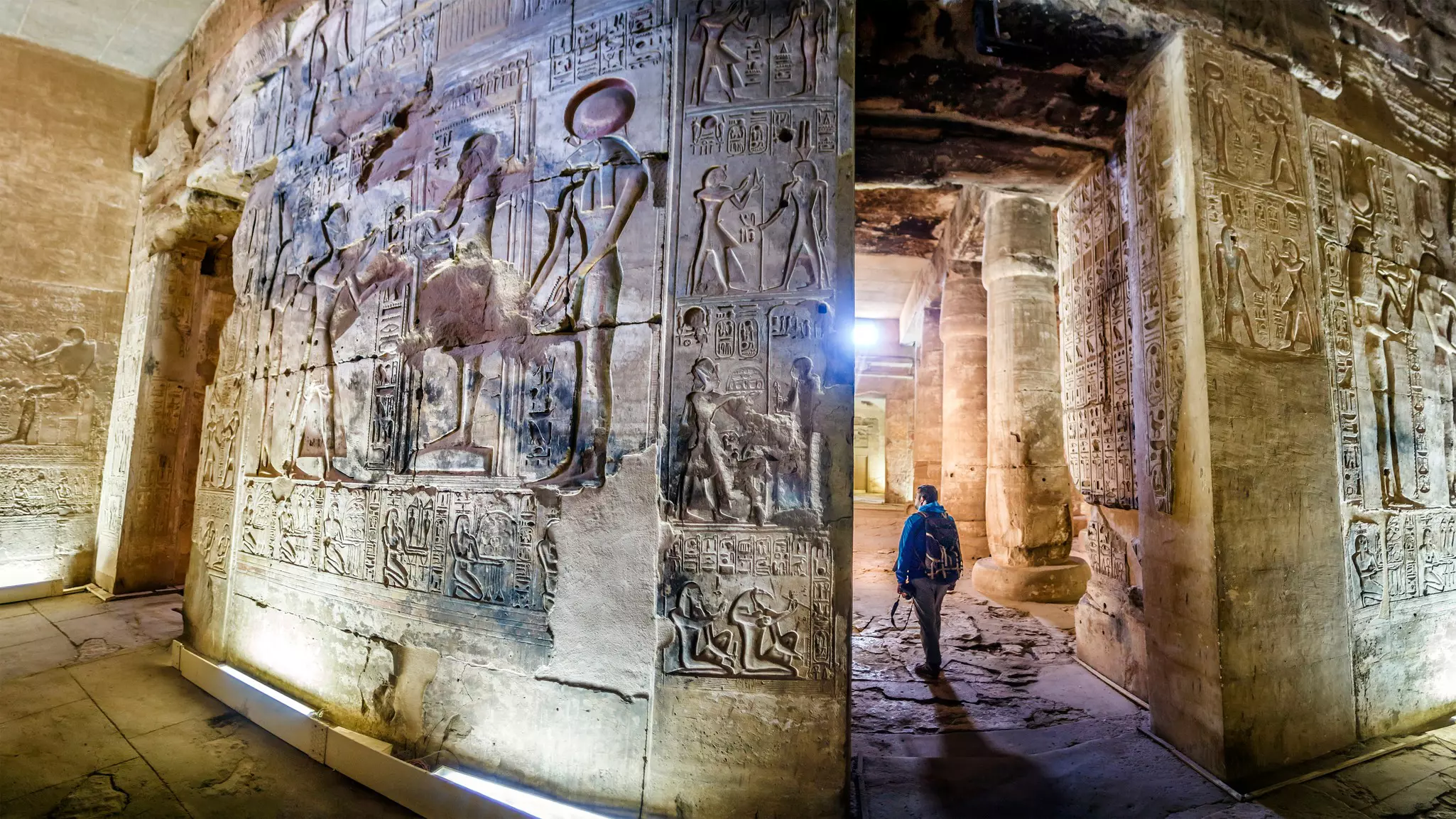 A person walks through an ancient temple with detailed carvings of people and writing covering all the walls.