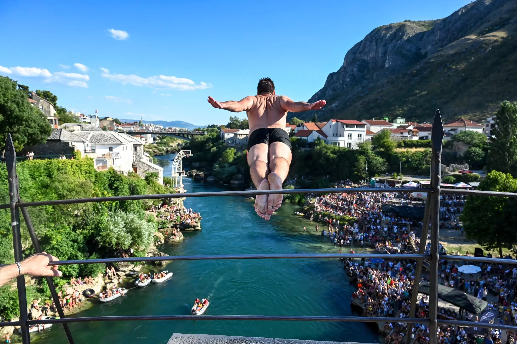 A bridge-jumper dives from a high point. The river and a crowd of onlookers are below them.