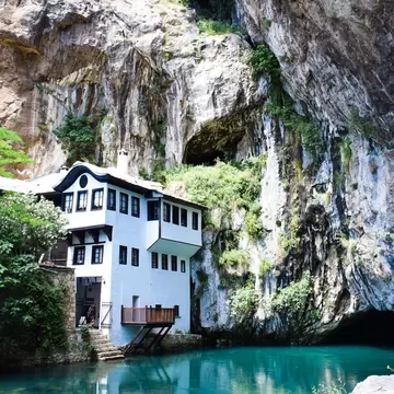 A white Ottoman-style house with dark window frames sits under a towering cliff and cave above turquoise water