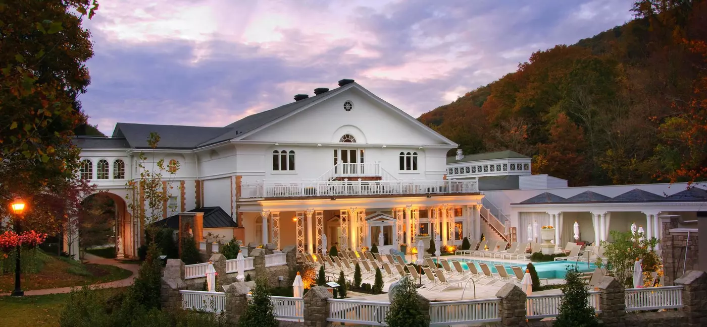A historic white building sits beside a pool with lawn chairs at dusk, with fall foliage and mountains in the background