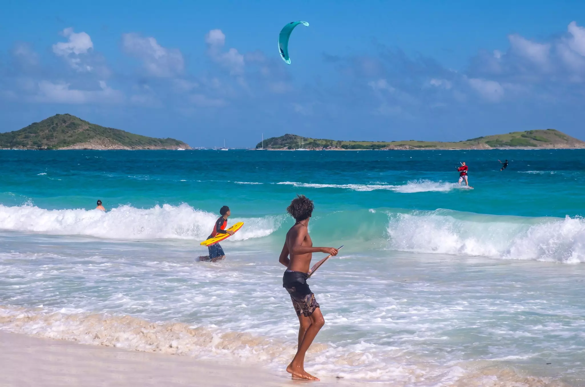 A young man watches surfers and kitesurfers in the water on a tropical beach.