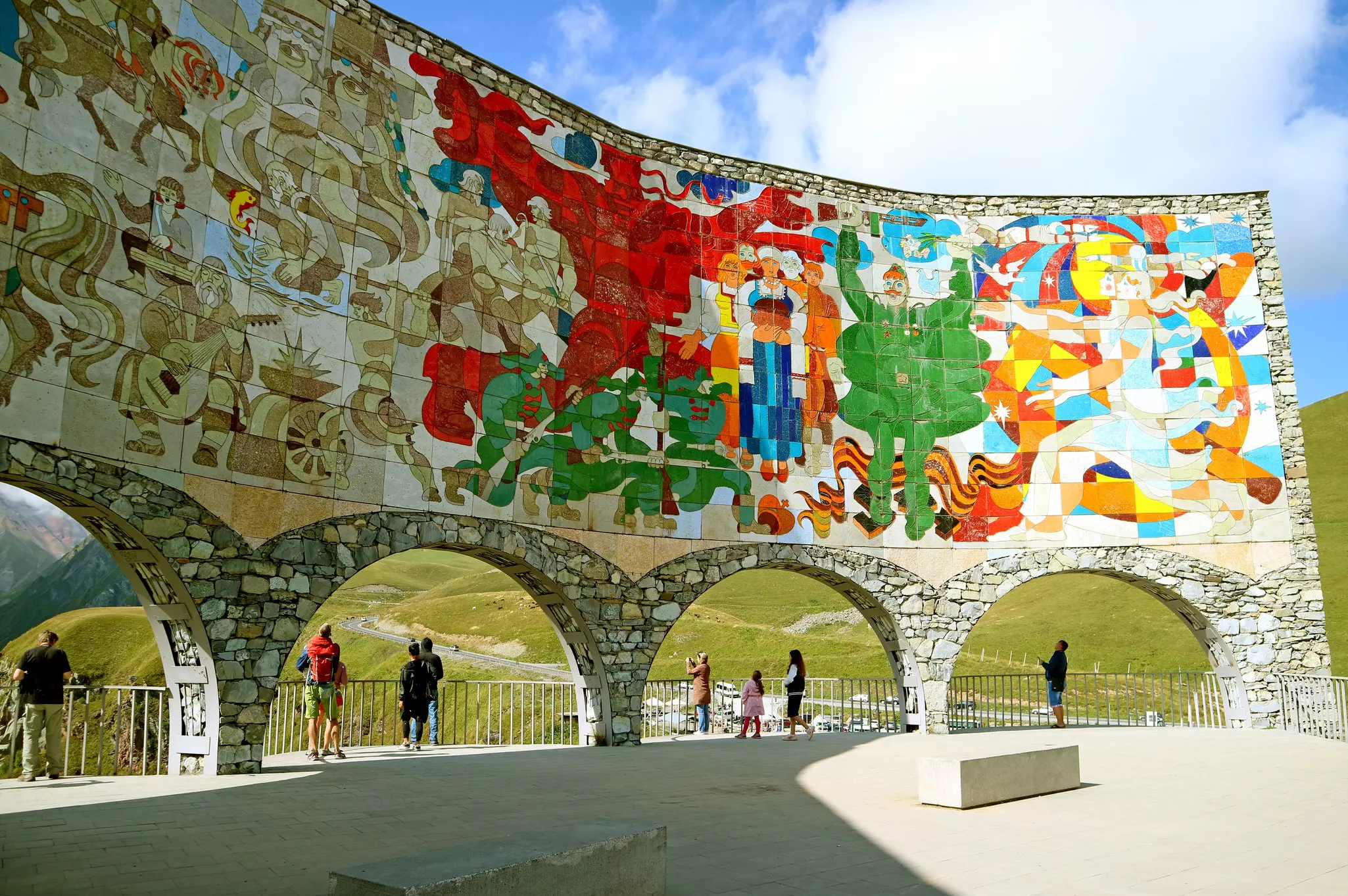 A colorful tiled mural on the interior of a curved structure built on a hillside. People stop to look out at a view over the valley.