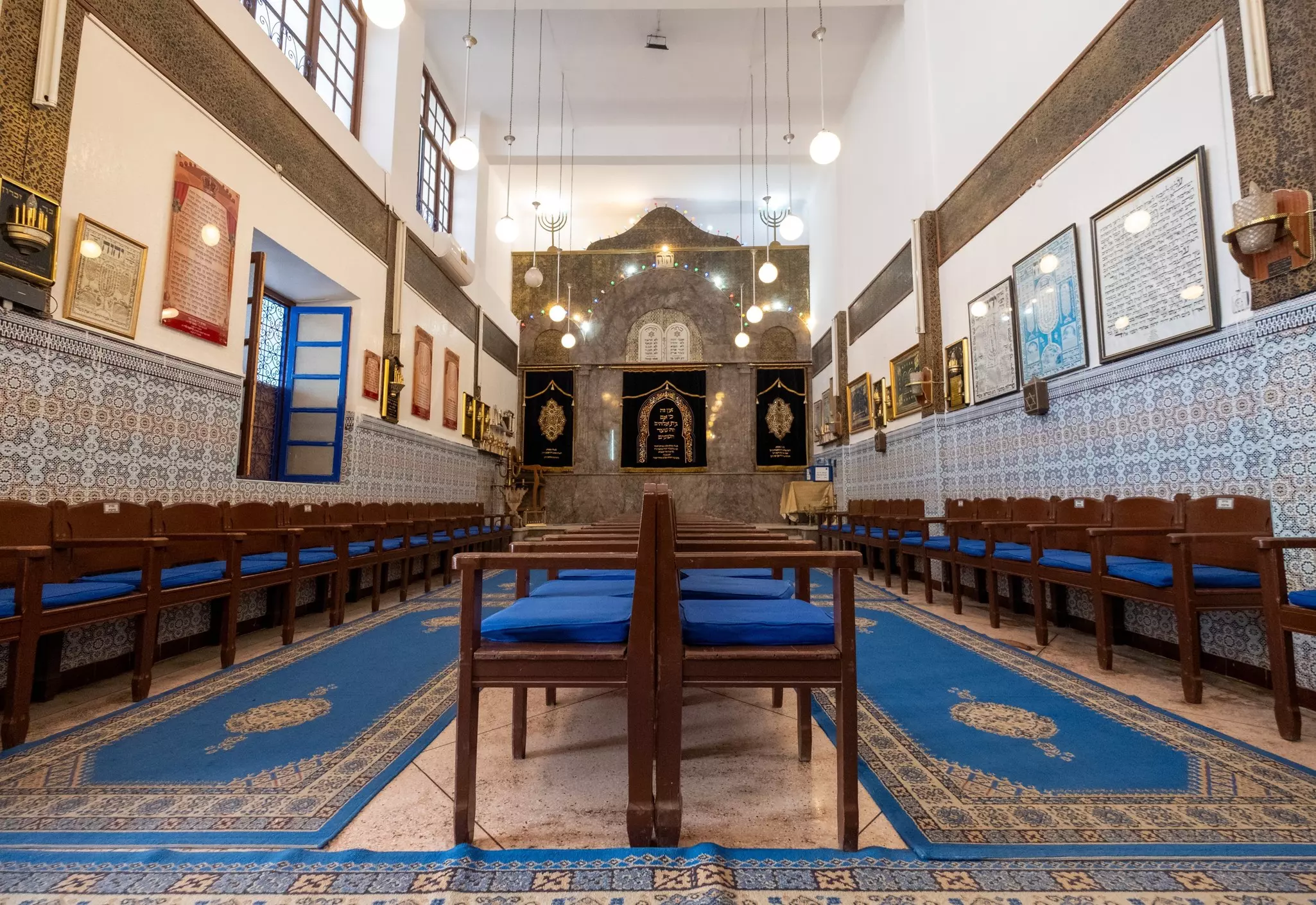 The prayer hall of a synagogue in Marrakesh has blue patterned carpets and tiled walls; wooden chairs with blue cushions are lined against the wall and in the center of the space.