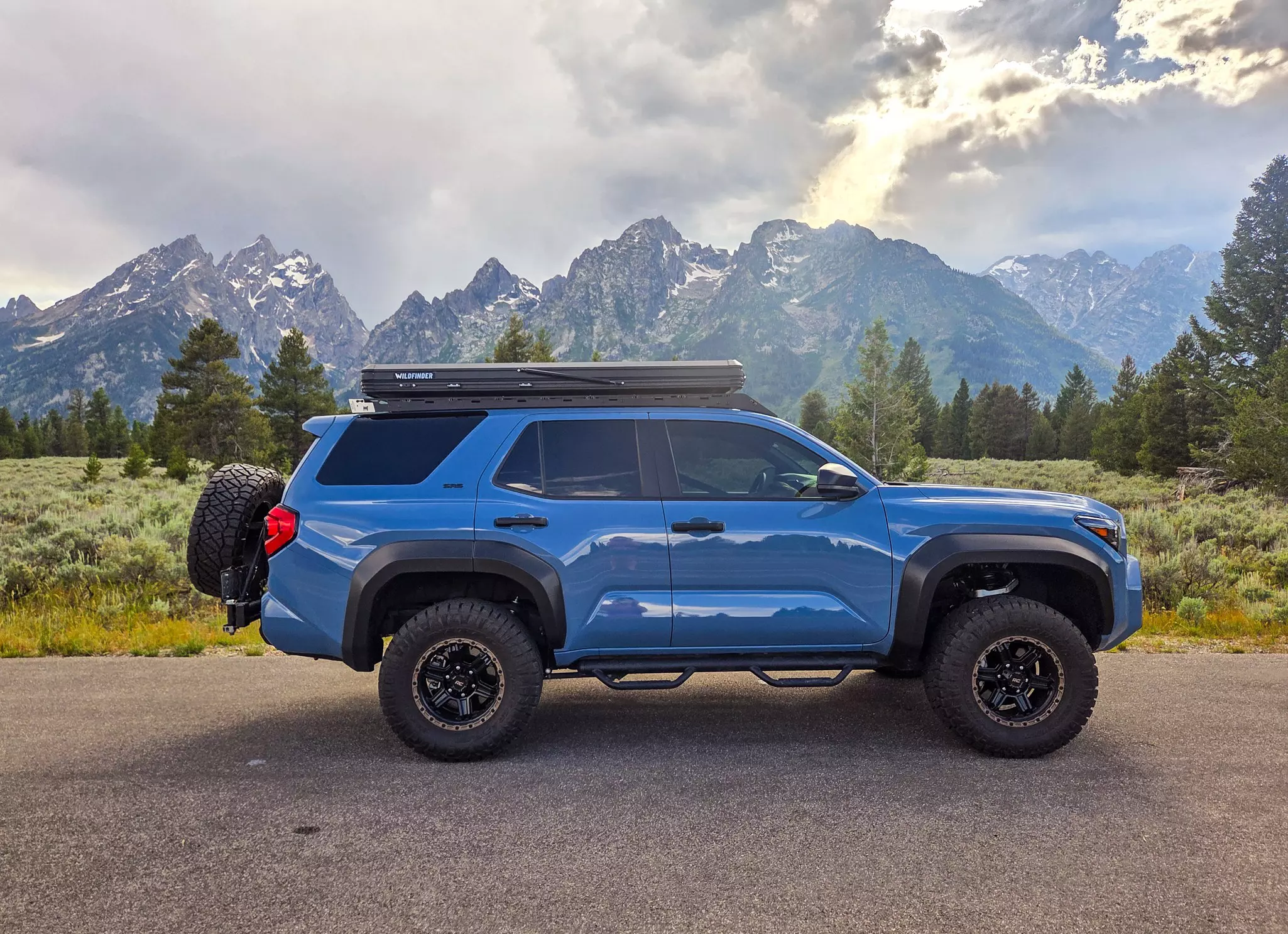 A blue SUV with a folded tent on its roof, in front of the Teton mountain range in Wyoming.