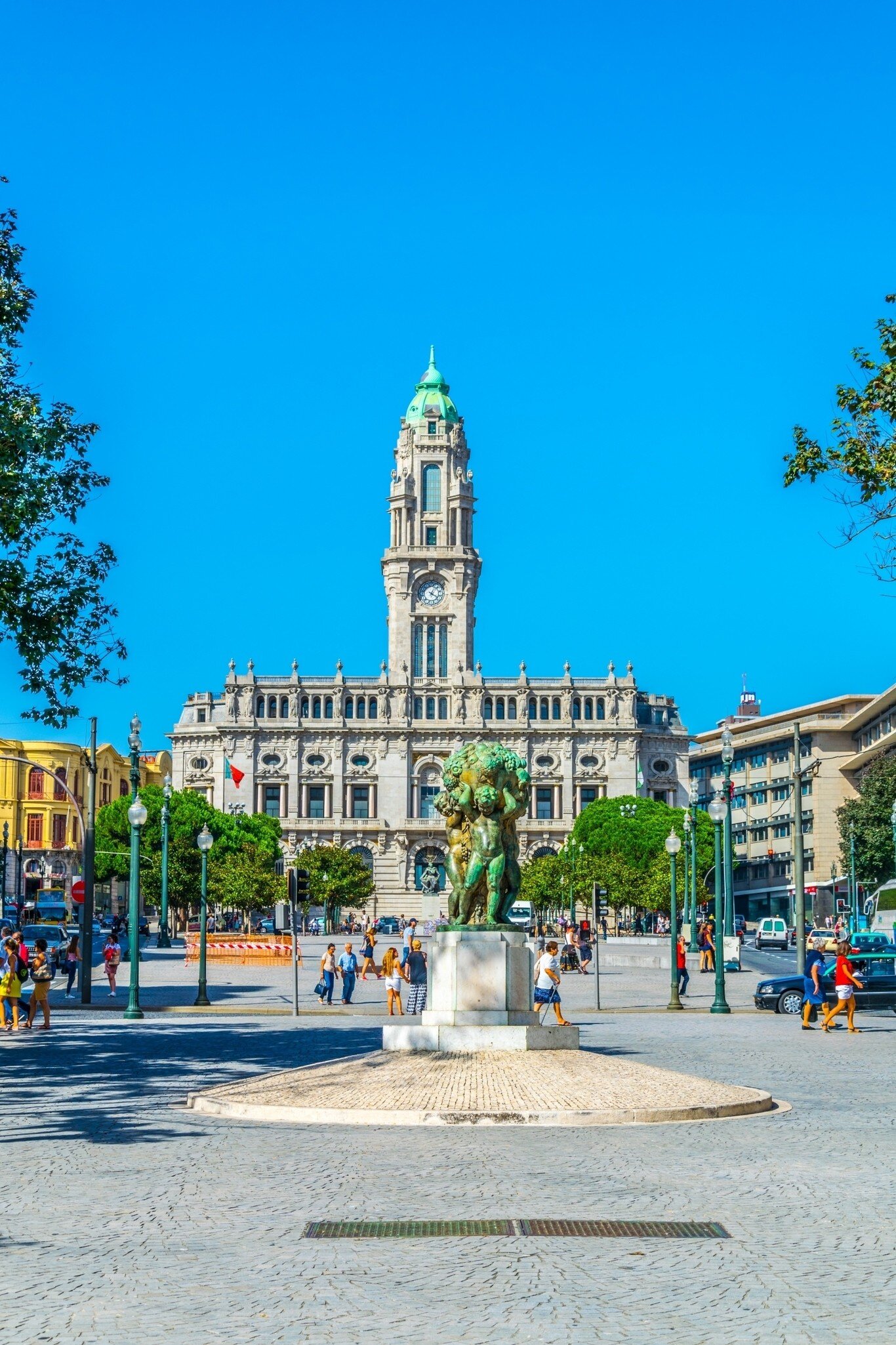 Granite city hall with a tall green-capped clock tower at the end of a wide plaza, a bronze statue and pedestrians, Porto.
