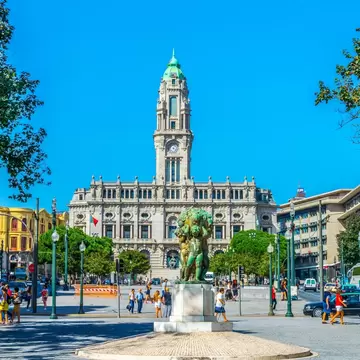 Granite city hall with a tall green-capped clock tower at the end of a wide plaza, a bronze statue and pedestrians, Porto.