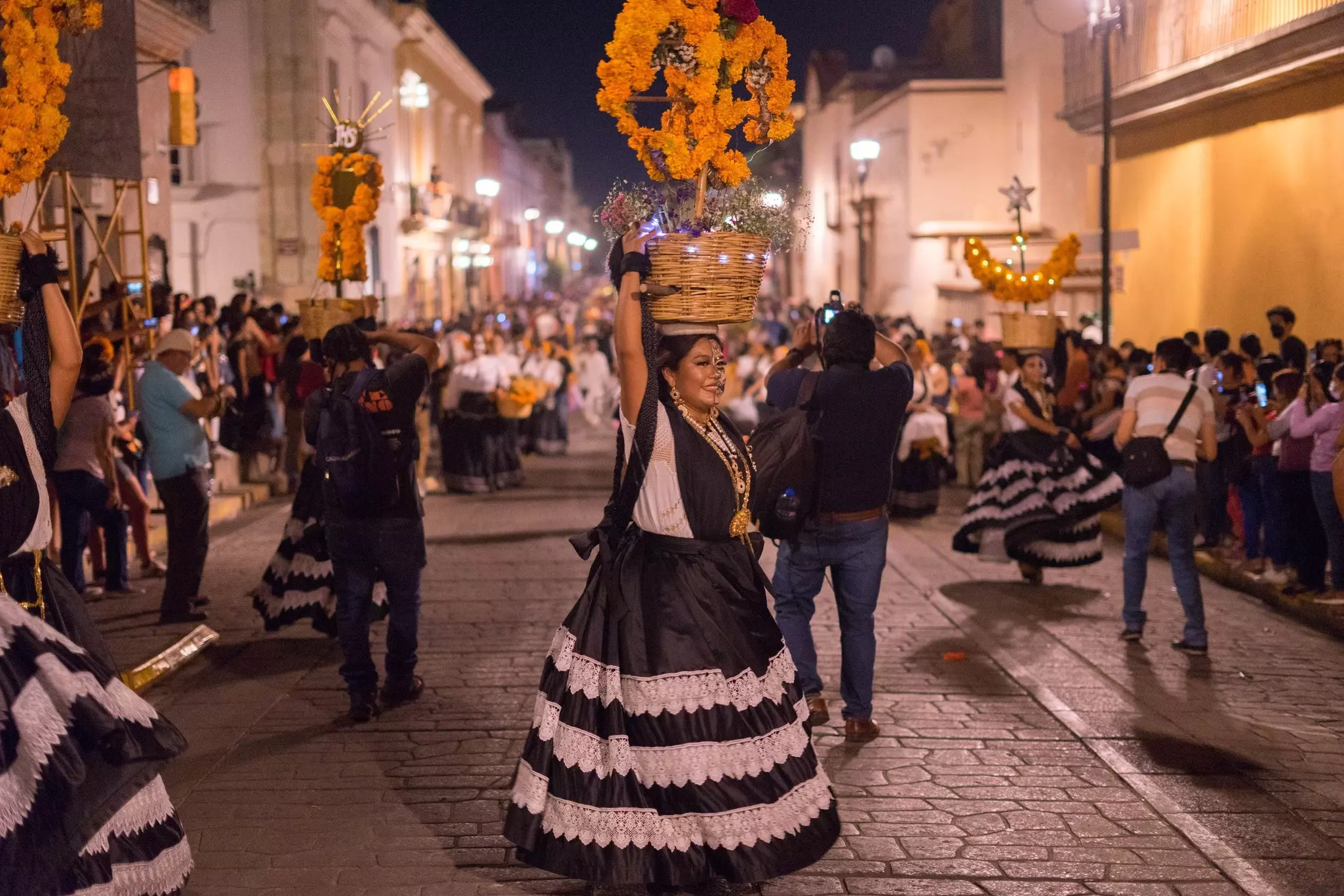 A woman in a black-and-white skirt and marigold headdress participates in a street parade in a city at night.
