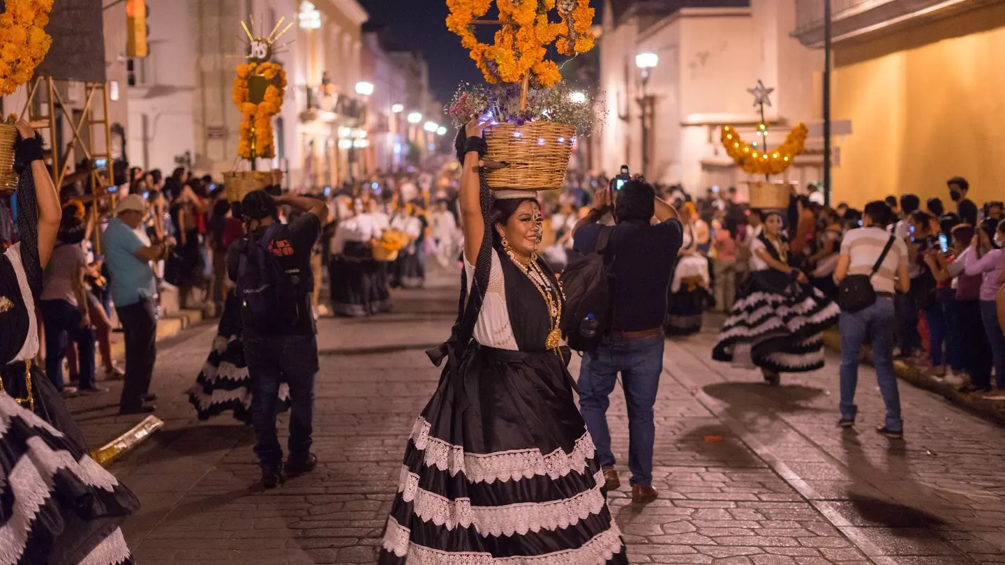 A woman in a black-and-white skirt and marigold headdress participates in a street parade in a city at night.