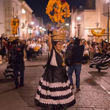 A woman in a black-and-white skirt and marigold headdress participates in a street parade in a city at night.
