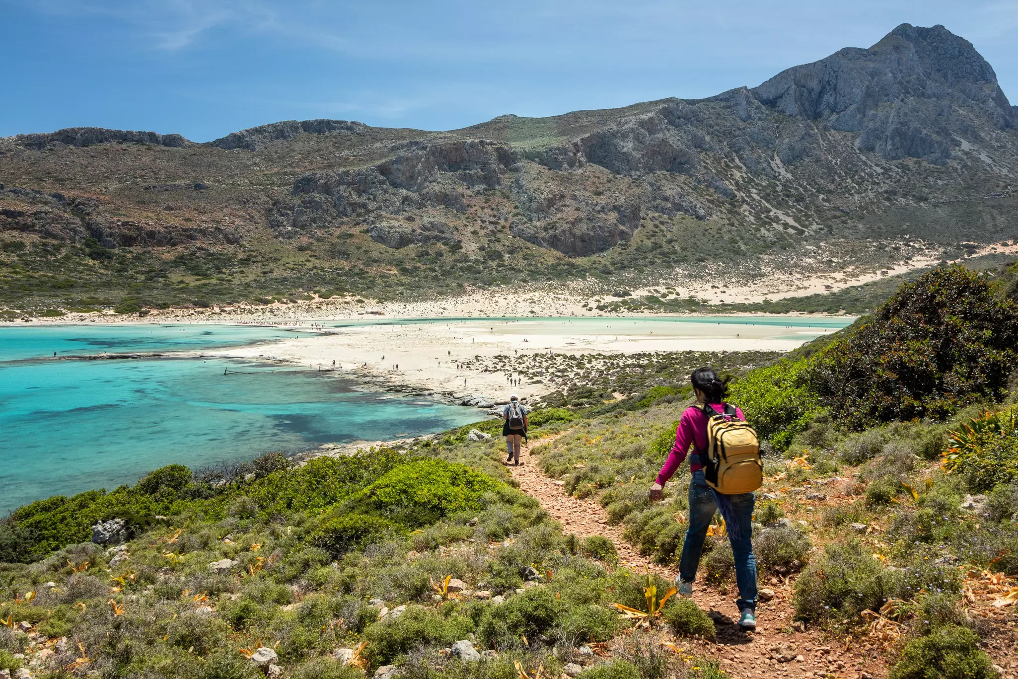 Two hikers follow a stony path down towards a beach lapped by a turquoise lagoon surrounded by hilly peaks.