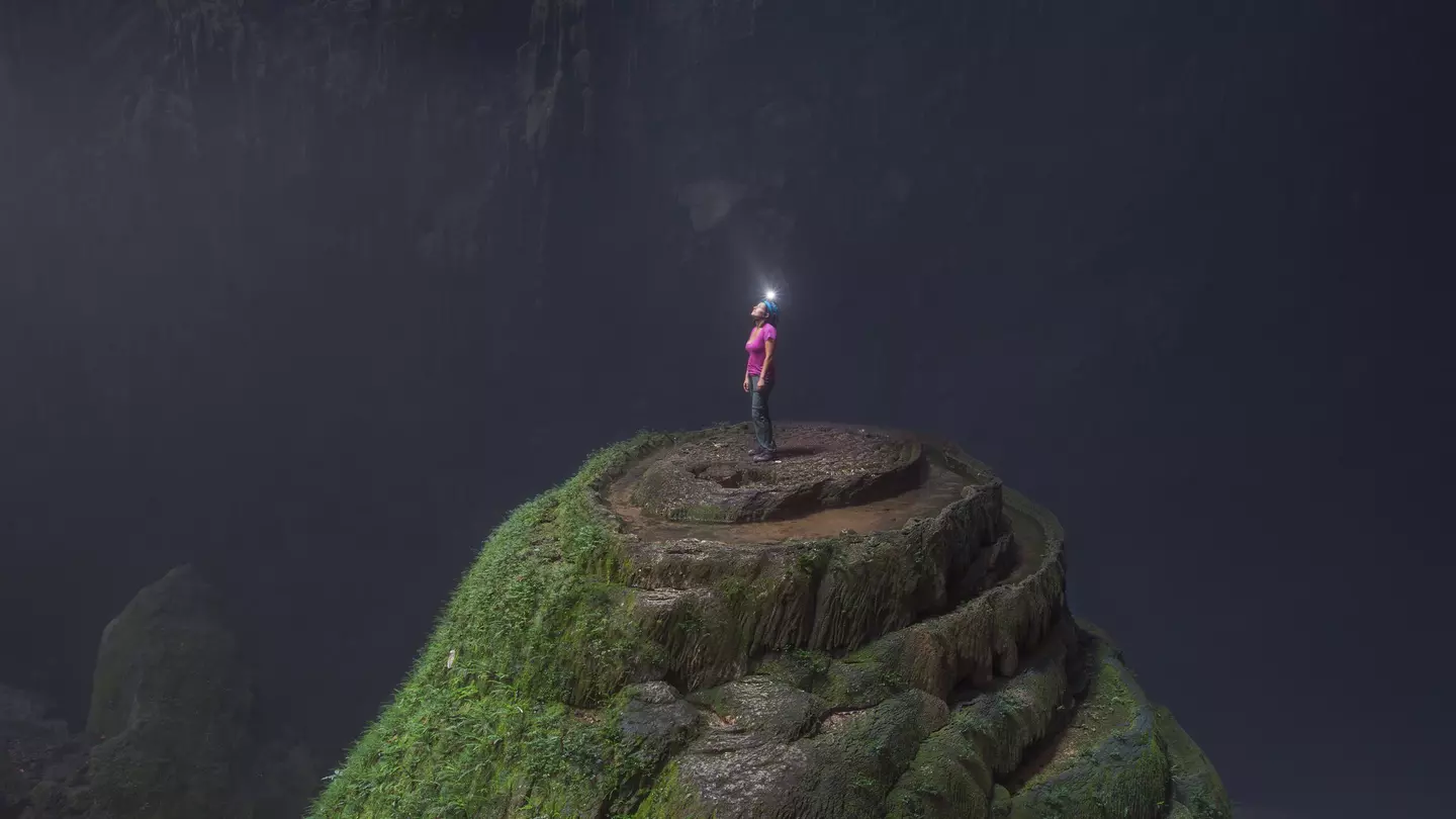 A woman wearing a helmet with a light stands at the top of a stalagmite inside Hang Son Doong cave.