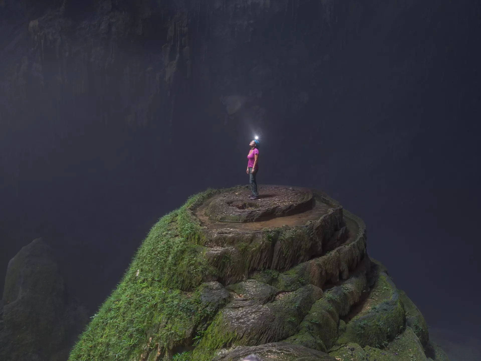 A woman wearing a helmet with a light stands at the top of a stalagmite inside Hang Son Doong cave.