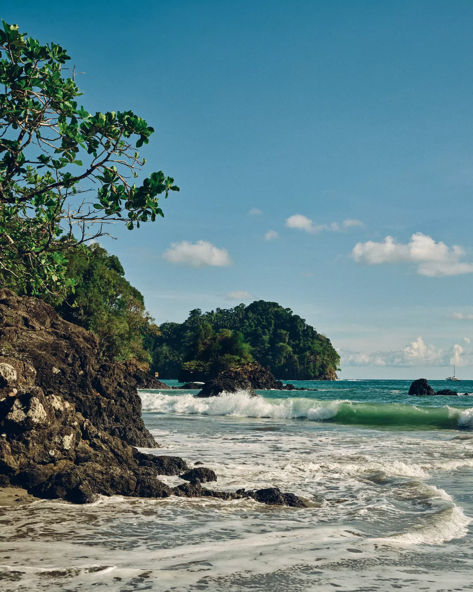 A large rock forms a cliff along the water of a beach in Costa Rica. Rocks jut out from the surf, trees cover the land that meets the sea. 
