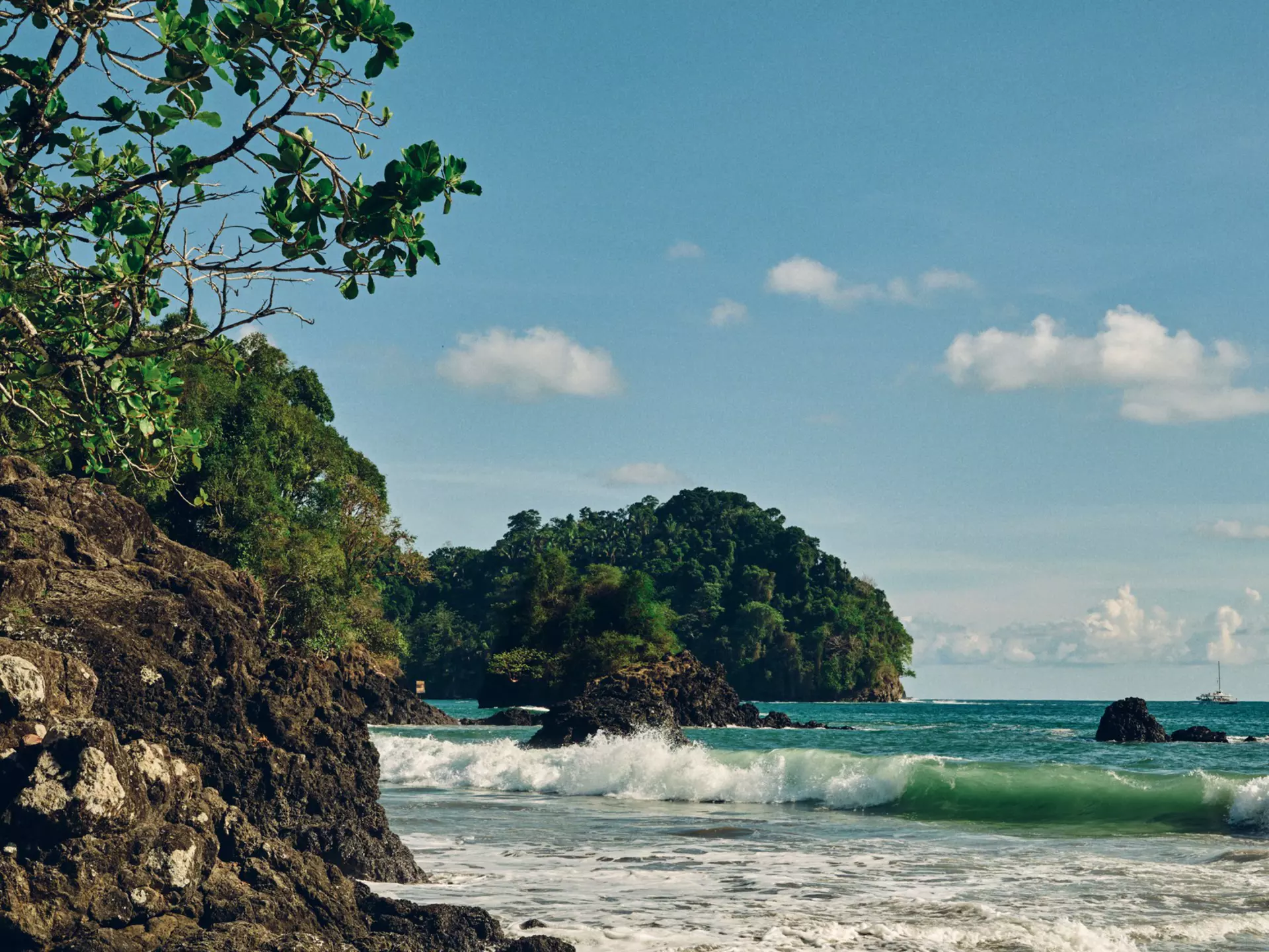 A large rock forms a cliff along the water of a beach in Costa Rica. Rocks jut out from the surf, trees cover the land that meets the sea. 