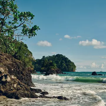 A large rock forms a cliff along the water of a beach in Costa Rica. Rocks jut out from the surf, trees cover the land that meets the sea. 