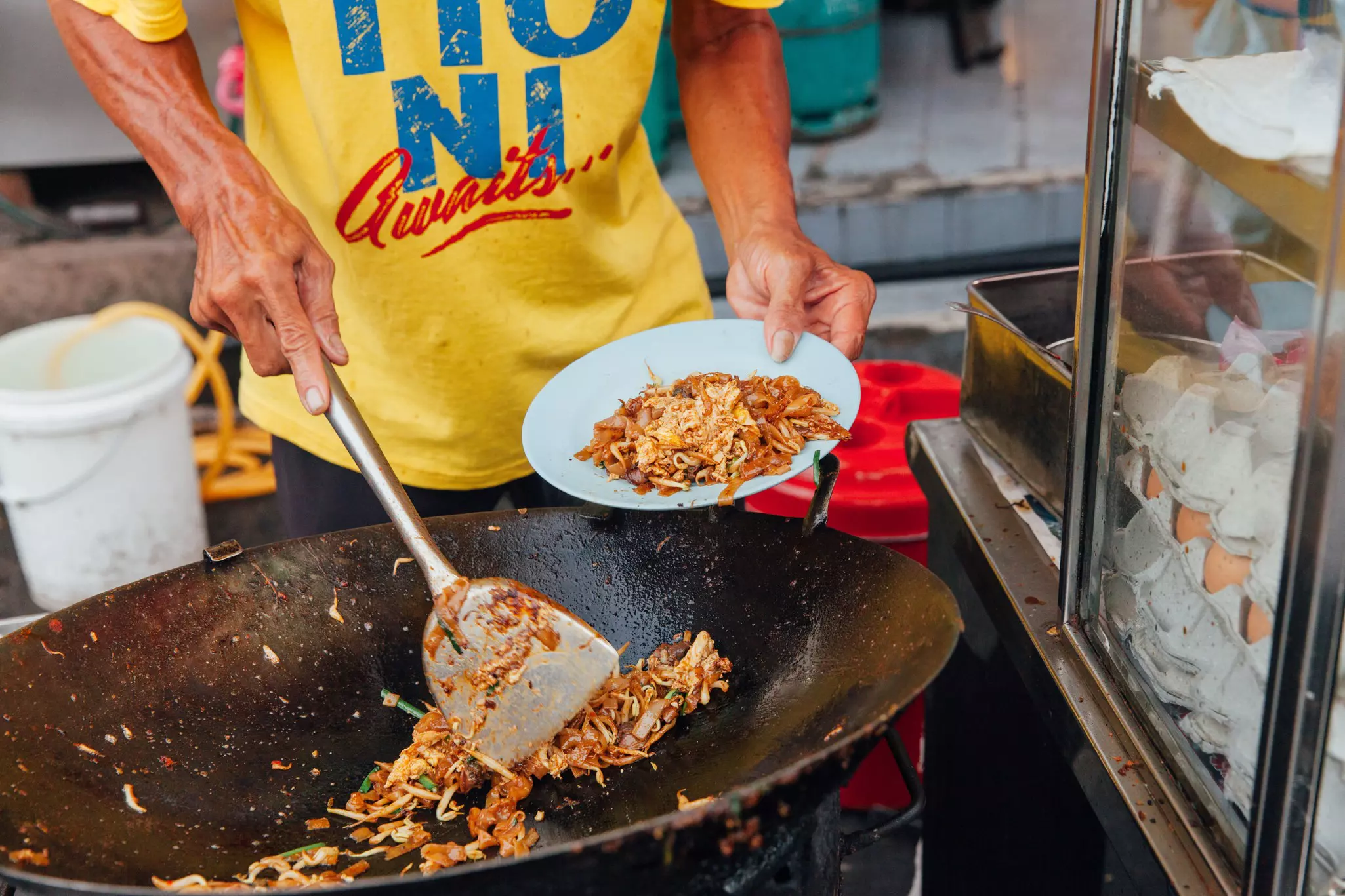 A person prepares a noodle dish in a wok at a street food market.
