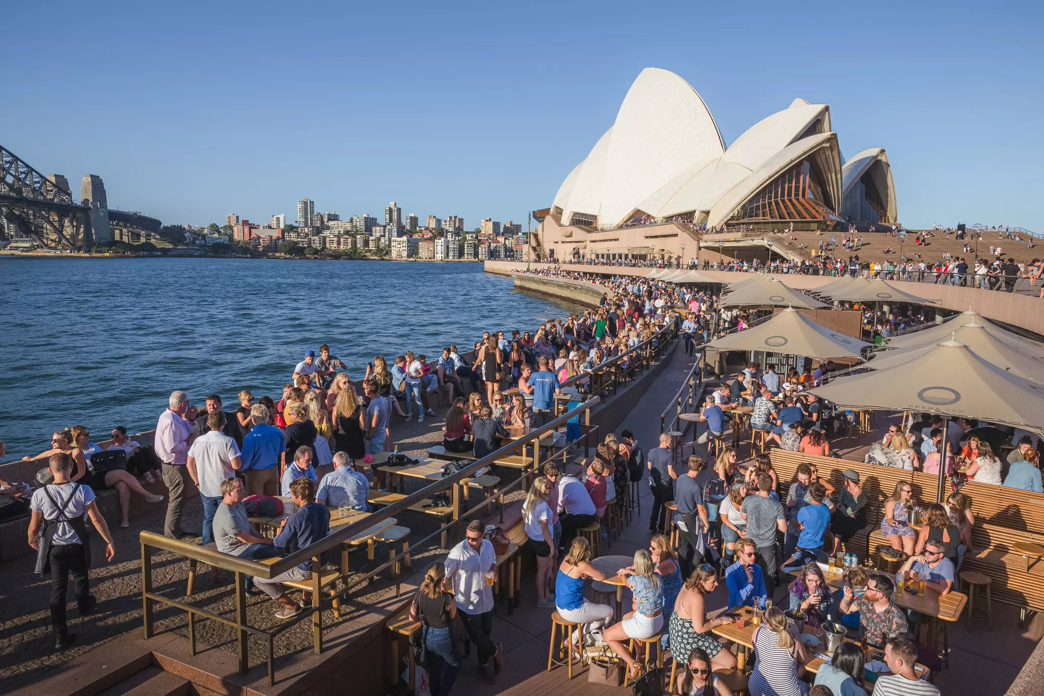People socialize at picnic tables on a rooftop near Sydney Harbor overlooking the opera house.