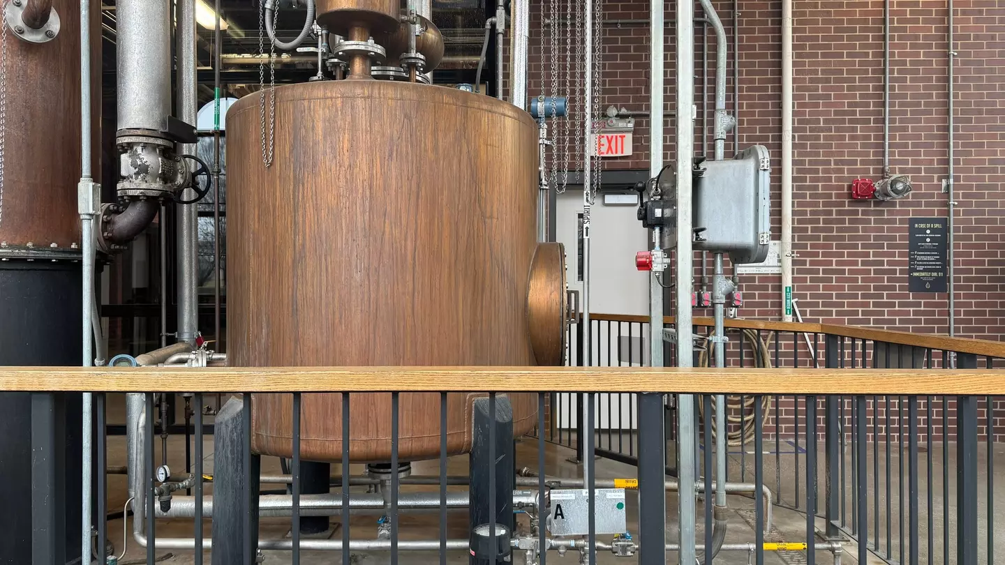 Close-up of distilling vat behind low metal barred enclosure and brick wall in the background.