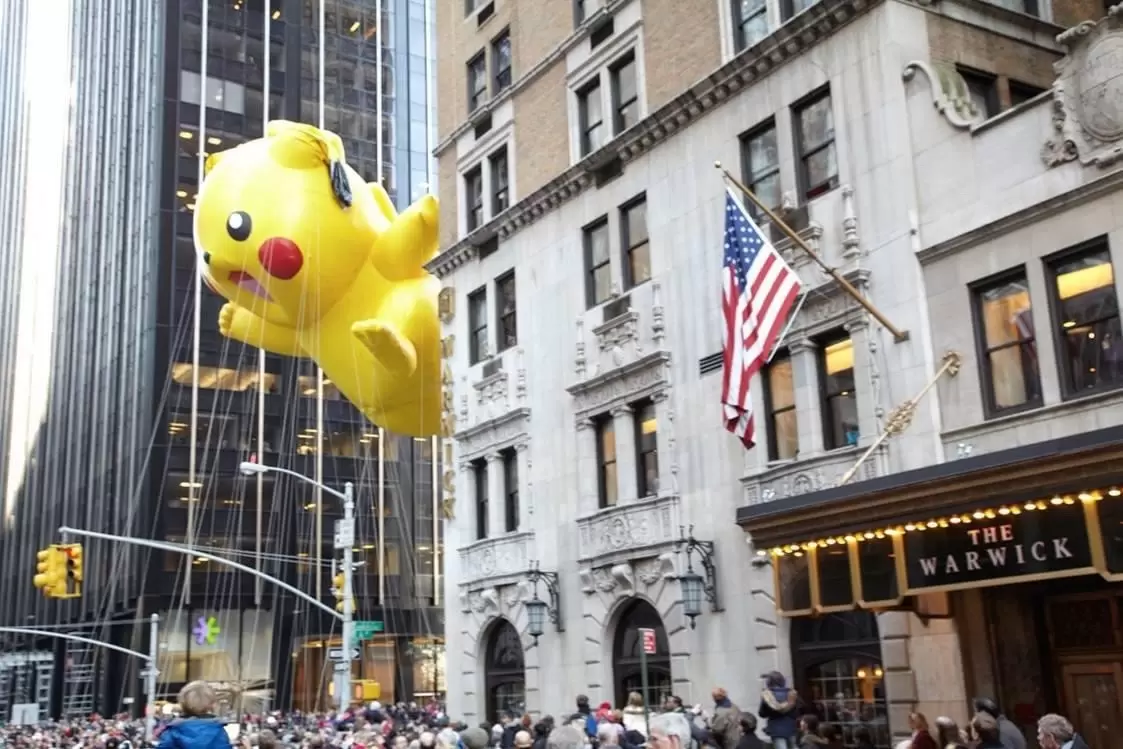A large yellow balloon character (Pikachu) passes a hotel as part of a parade: Warwick New York Hotel.