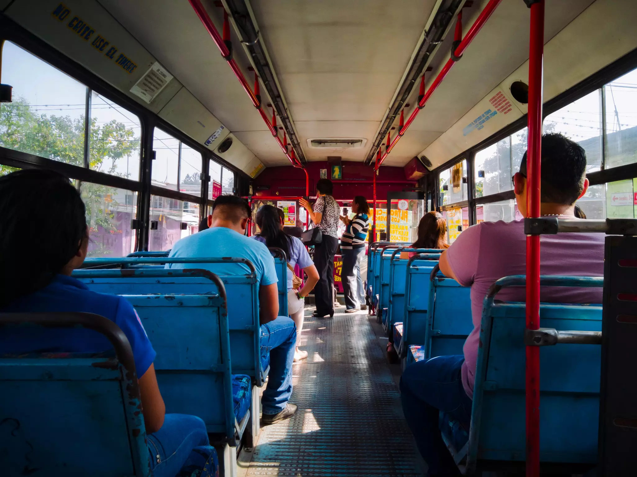 The interior of a public transportation bus seen from the back, with people sitting on blue seats on either side of an aisle. A few people stand at the front of the bus holding rails.