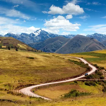 A car weaves down a road backed by mountains on a sunny day.