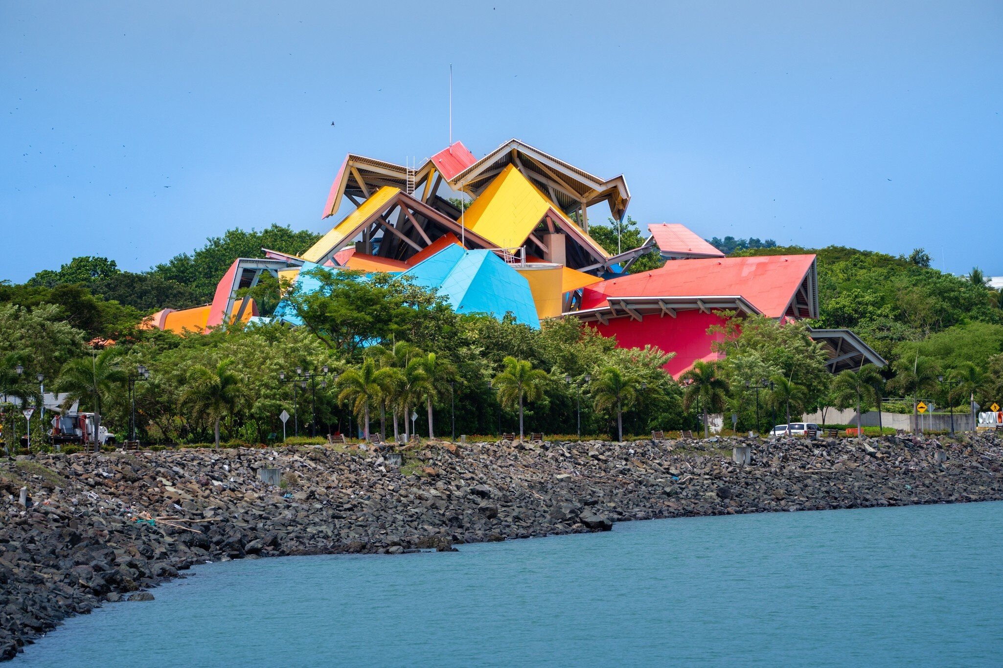 Angular red, yellow and blue roof panels above palm trees, a rocky seawall and calm water in front.