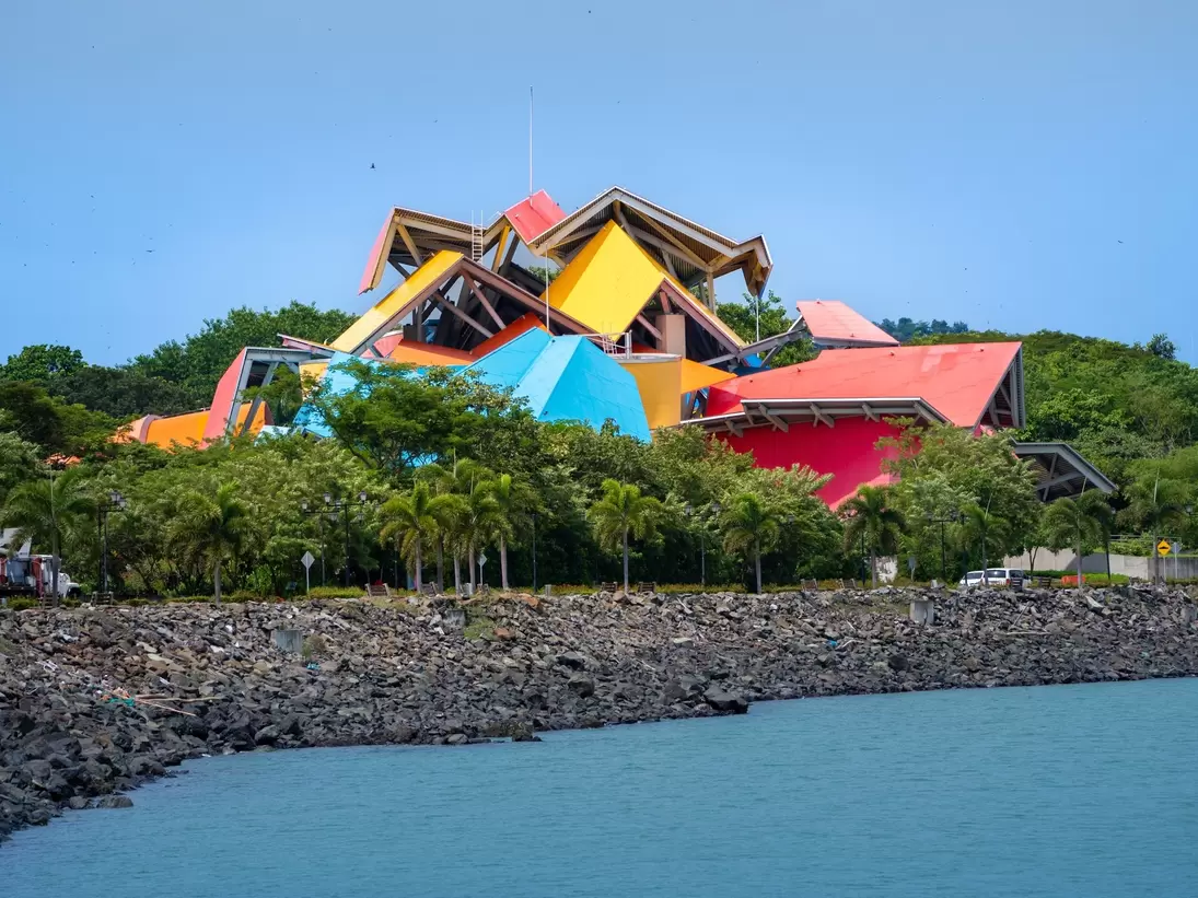 Angular red, yellow and blue roof panels above palm trees, a rocky seawall and calm water in front.