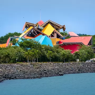Angular red, yellow and blue roof panels above palm trees, a rocky seawall and calm water in front.
