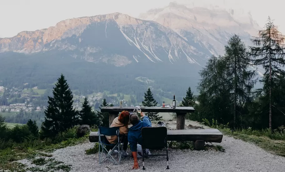 Couple at a wooden picnic table with wine, looking across a valley town and pine forest to rocky Dolomite peaks
