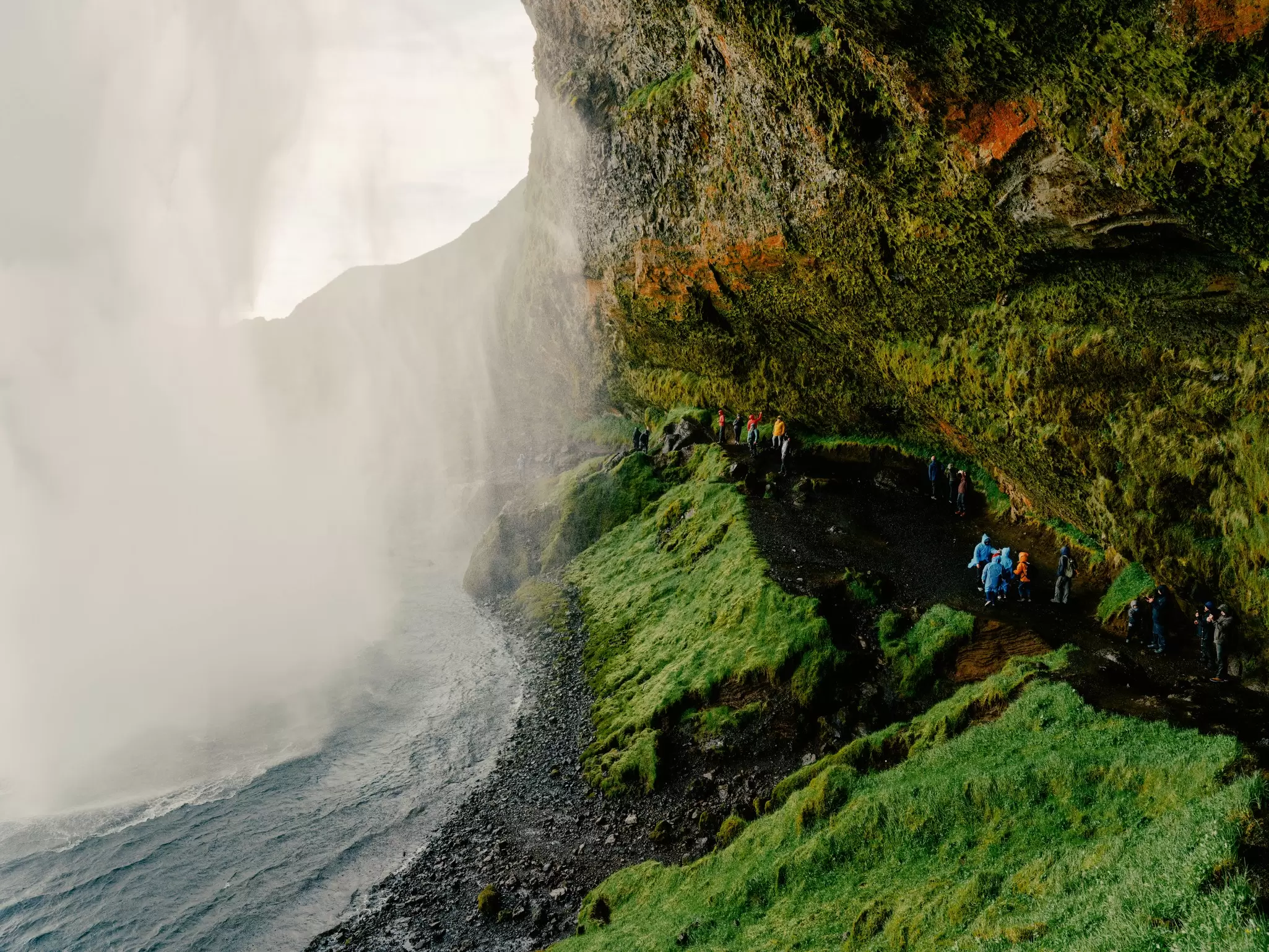 People walk on a ledge under a green hillside toward the mist of a waterfall.