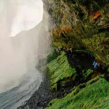 People walk on a ledge under a green hillside toward the mist of a waterfall.