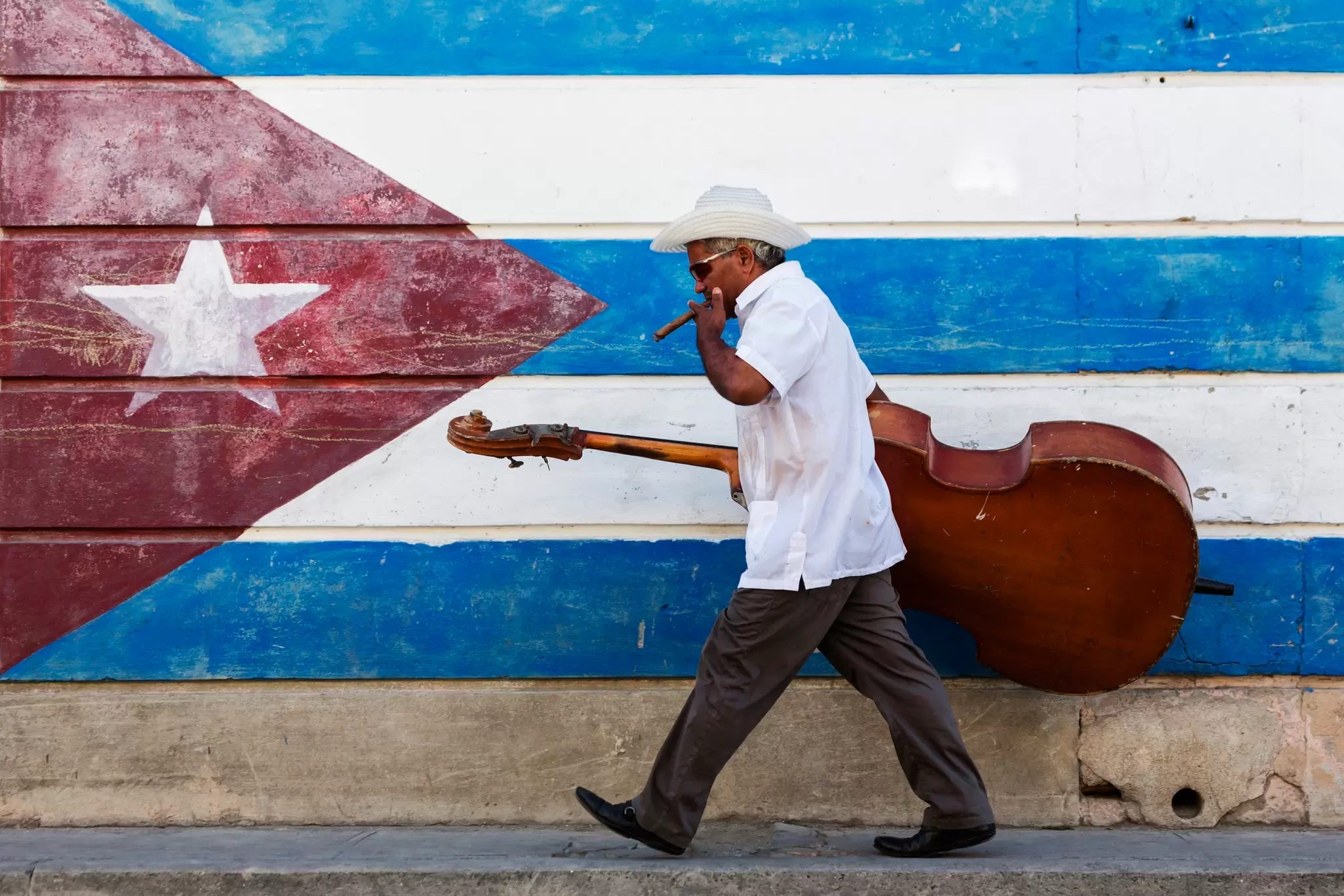 A man carrying a large bass instrument smokes a cigar as he walks by a Cuban flag painted on a wall